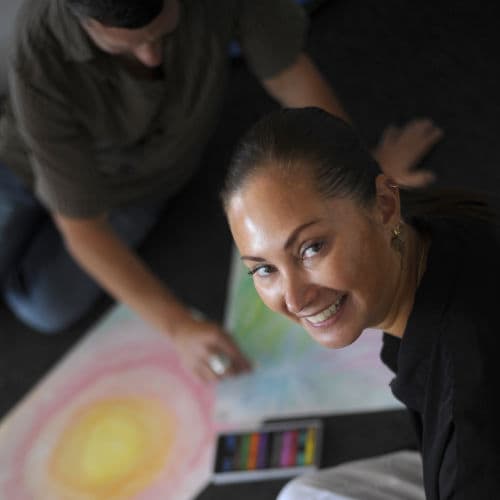 A smiling student studying Advanced Diploma of Transpersonal Therapy sitting on the floor with artwork.