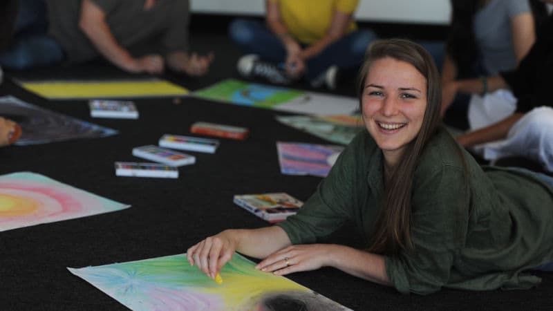 A smiling student studying Advanced Diploma of Transpersonal Therapy lying on the floor with artwork.