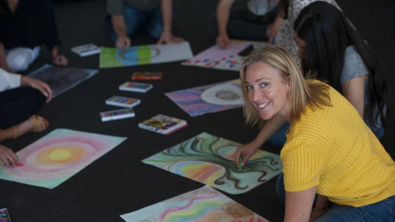 A smiling Advanced Diploma of Transpersonal Therapy student sitting in a circle of students with their artwork.