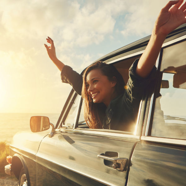 A smiling woman with her head and outstretched arms out of the passenger side of a vehicle parked near the ocean.