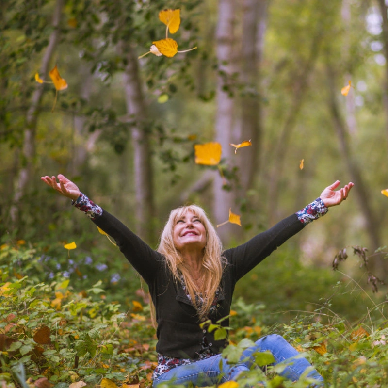 A woman sites in a forest surrounded by flowers with her arms wide and hands raised in the air.